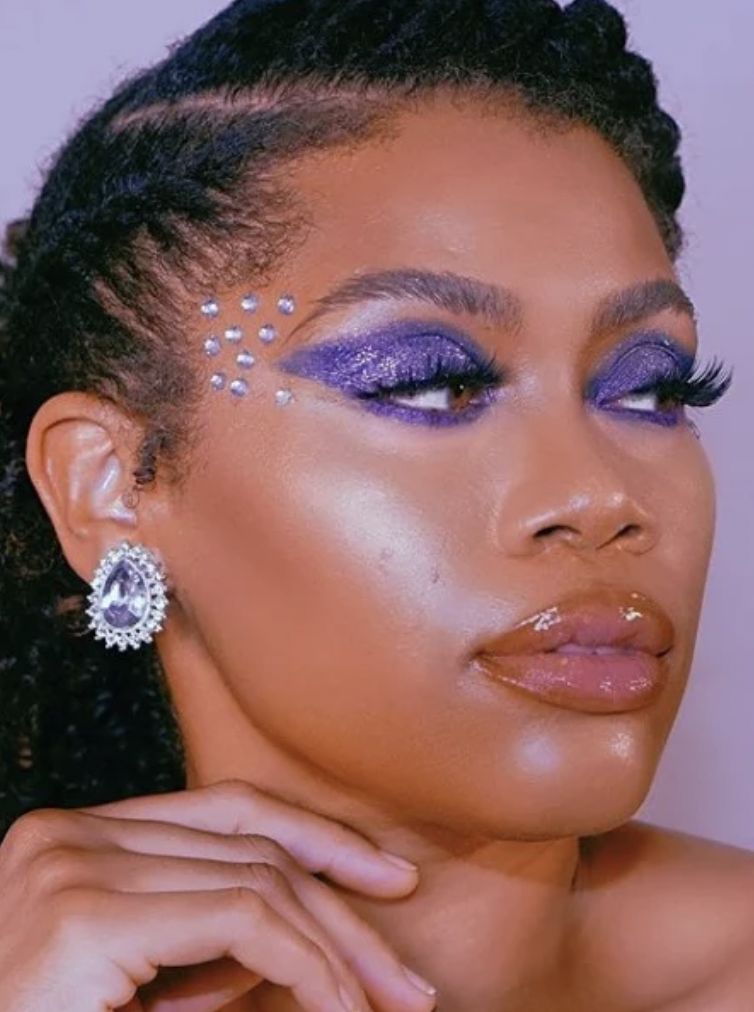 A woman with bright purple eyeshadow blended outwards and silver rhinestones applied to her temples. 