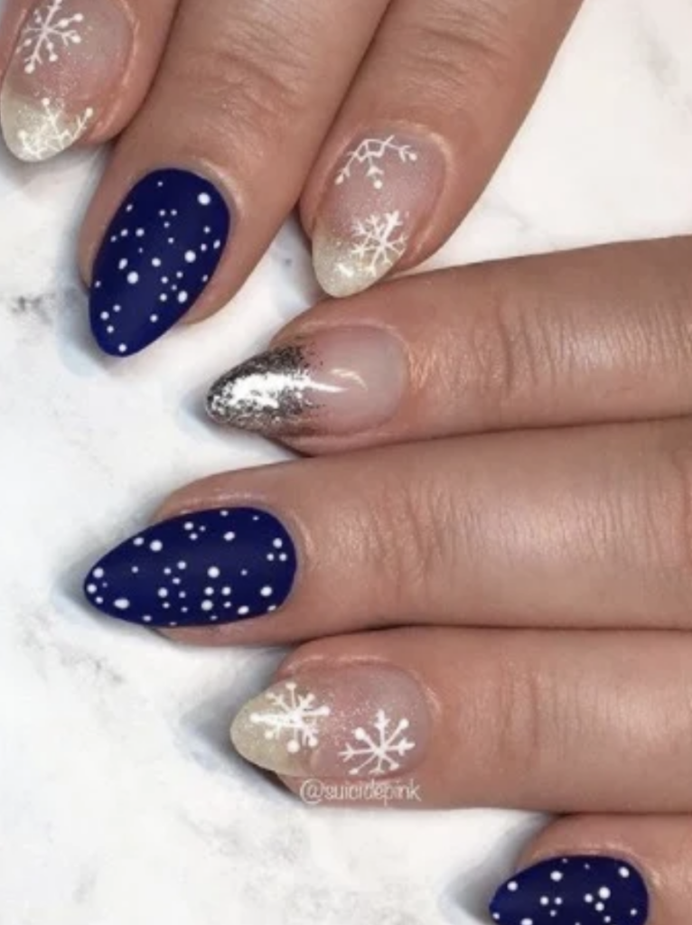 Mid length almond shaped winter themed nails with decals of white snowflakes, silver foil and a blue base with small white dots. 
