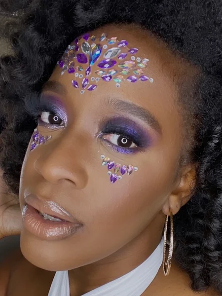 A woman with purple eyeshadow and blue, green, silver, and purple jewels applied to her forehead and under eye areas. 