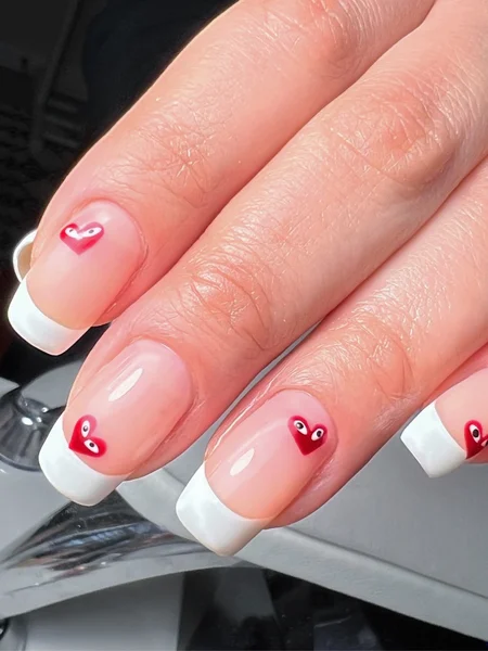 Traditional french manicure with red heart decals that have eyes drawn on them. 