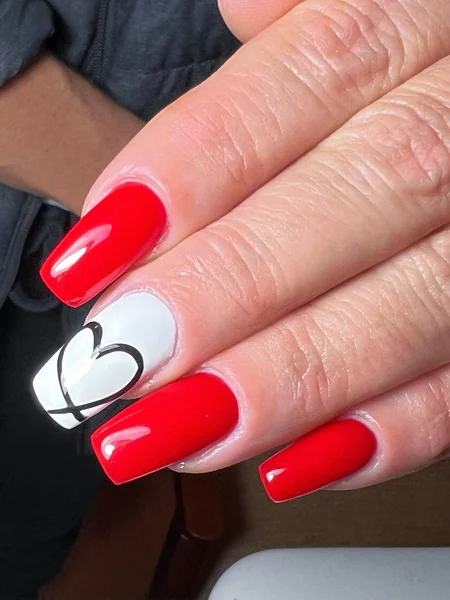 Red nails with one white nail that has a simple black heart decal. 