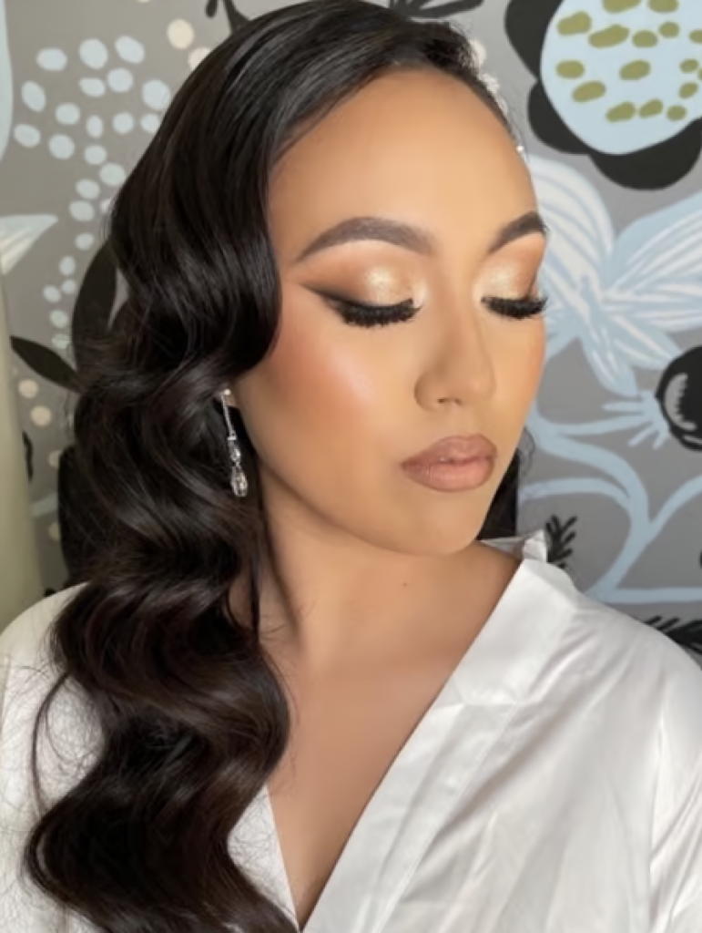 A woman with black, curled hair parted to the side with soft glam makeup, black cat eyeliner, and nude lips. 