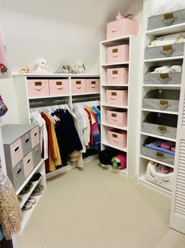 A kid's walk in closet organized with pink bins and clothes on hangers that are sorted by color. 