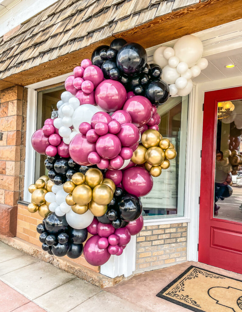 A white, pink, black and gold balloon garland outside of a storefront. 
