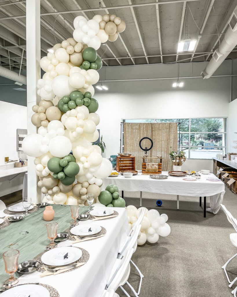 A large white, cream, beige and sage colored moon shaped balloon garland in between two banquet tables for an influencer gifting event. 