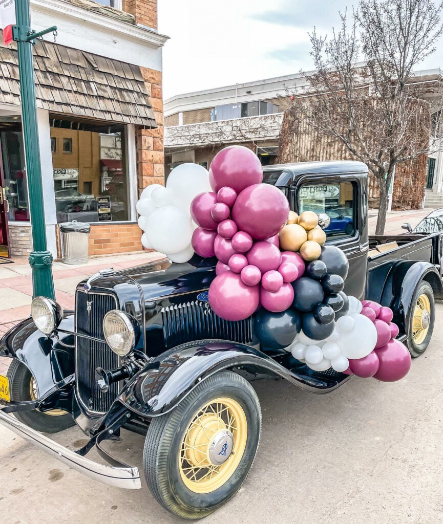A white, pink, black and gold balloon garland on top of a vintage car 