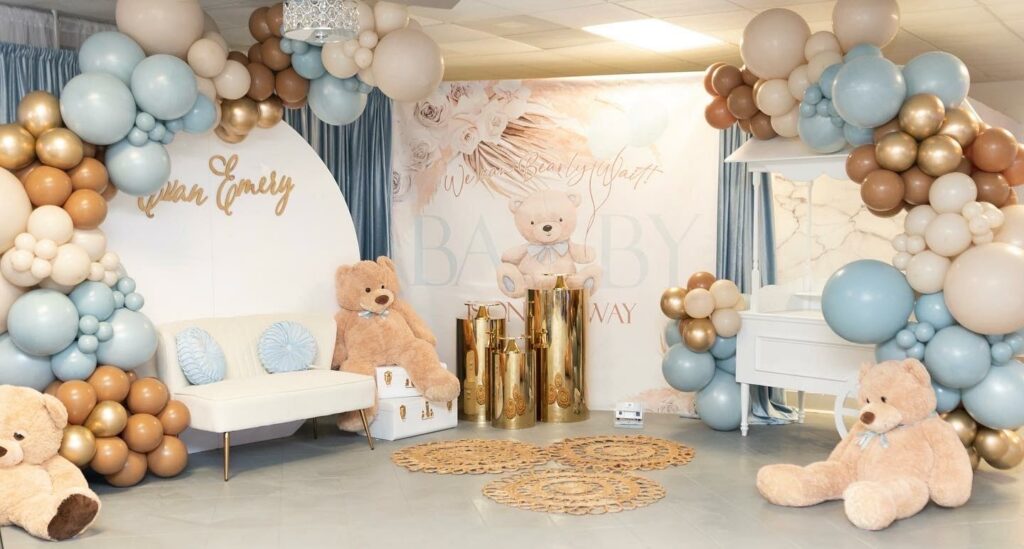 Multiple gold, white and blue balloon garlands scattered around a couch and bear stuffed animals for a baby shower. 