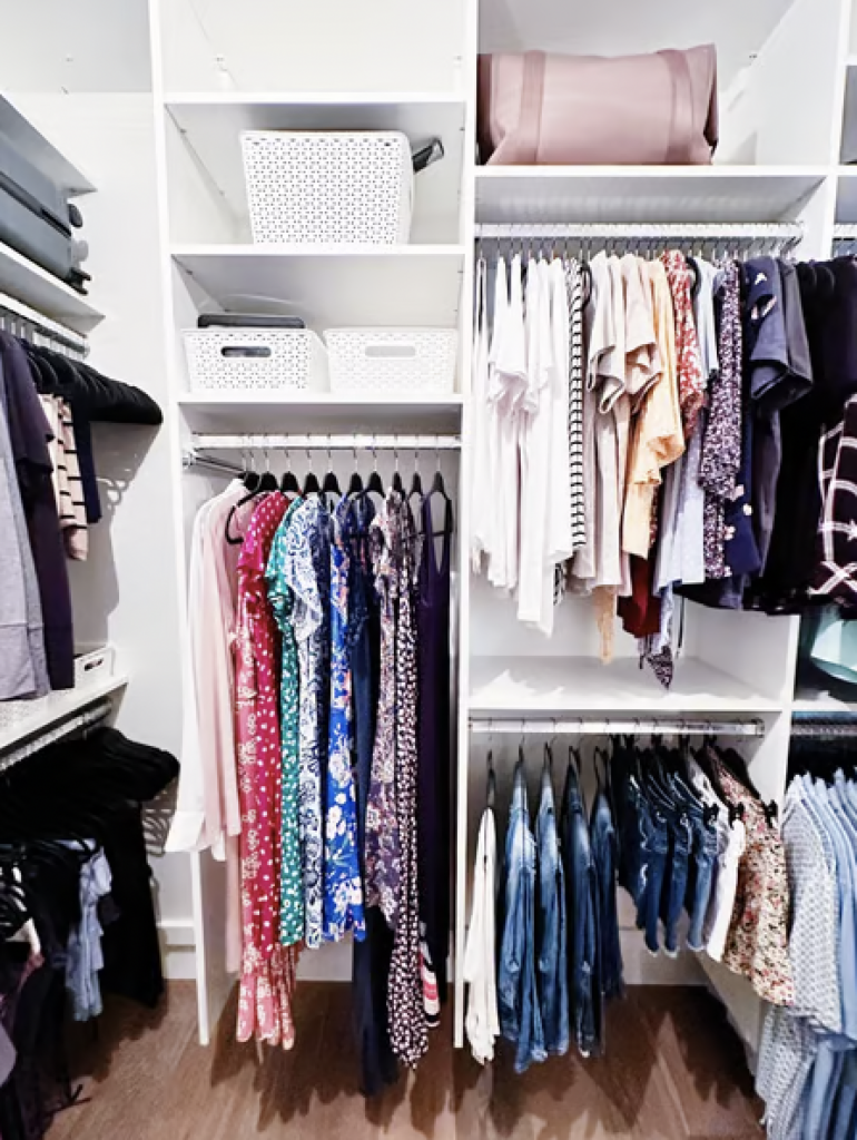 A professionally organized closet with clothes hanging neatly on hangers and white storage bins. 