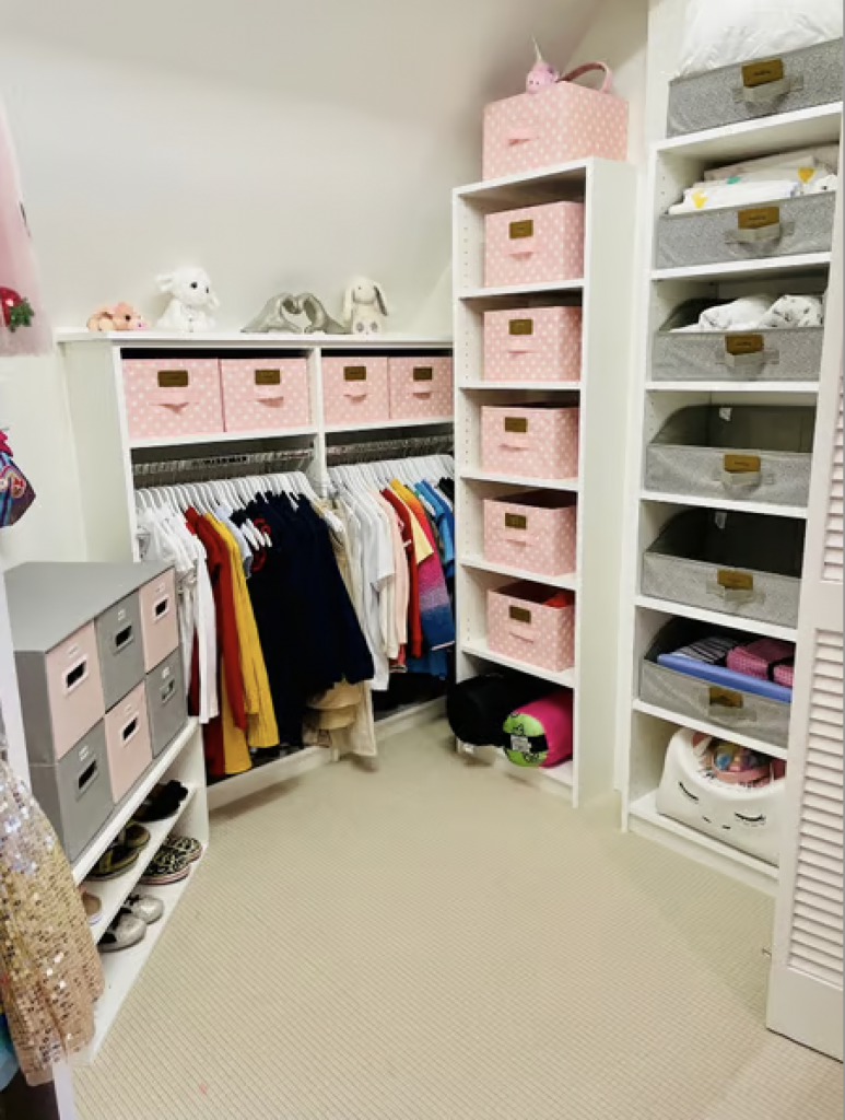 A kid's walk in closet that has been organized with pink and grey storage bins and the clothes are color coordinated on hangers. 