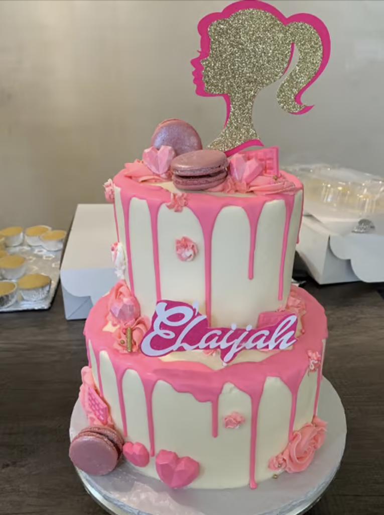 A two-tiered white cake with hot pink icing dripping down the edges, a barbie decal, and pink macaroons. 