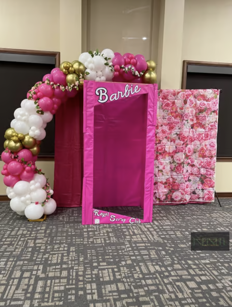 A life size, pink barbie box with a pink, gold and white balloon garland. 