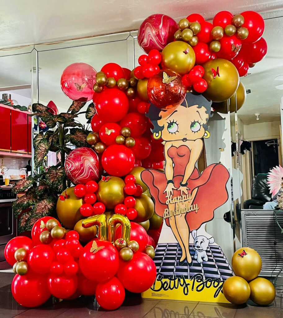 A red and gold balloon garland around a Betty Boop cardboard cutout for a 70-year-old's birthday party. 