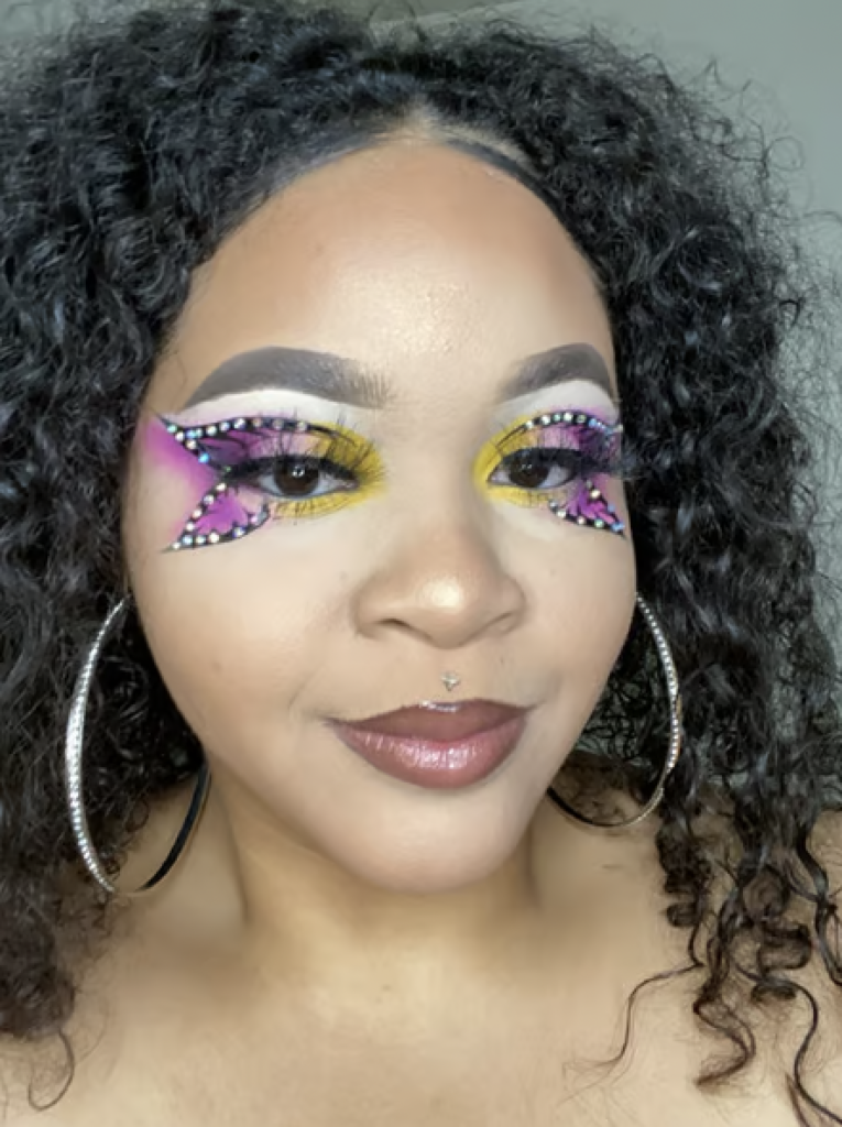 Eyeshadow look with yellow inner corners, white on her upper eyelid, and purple on her outer corners creating a butterfly look with a black outline and rhinestones. 