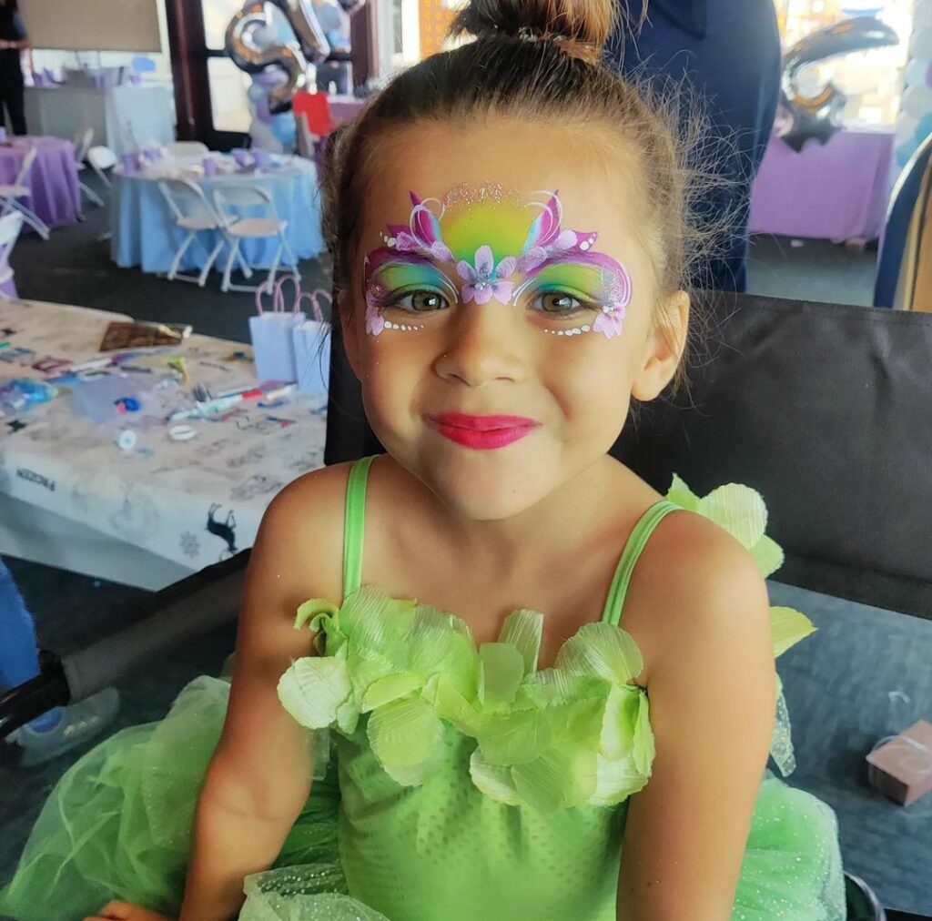Green, purple, pink and blue floral princess face paint with a flower in the middle. 