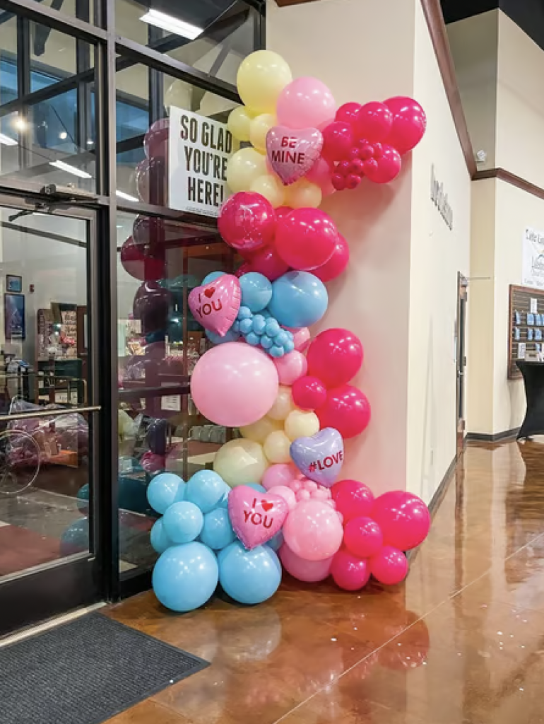 Pink, yellow and blue balloons in the corner of a public space with decorative heart balloons that say "I Love You" and "Be Mine" for Valentine's Day. 