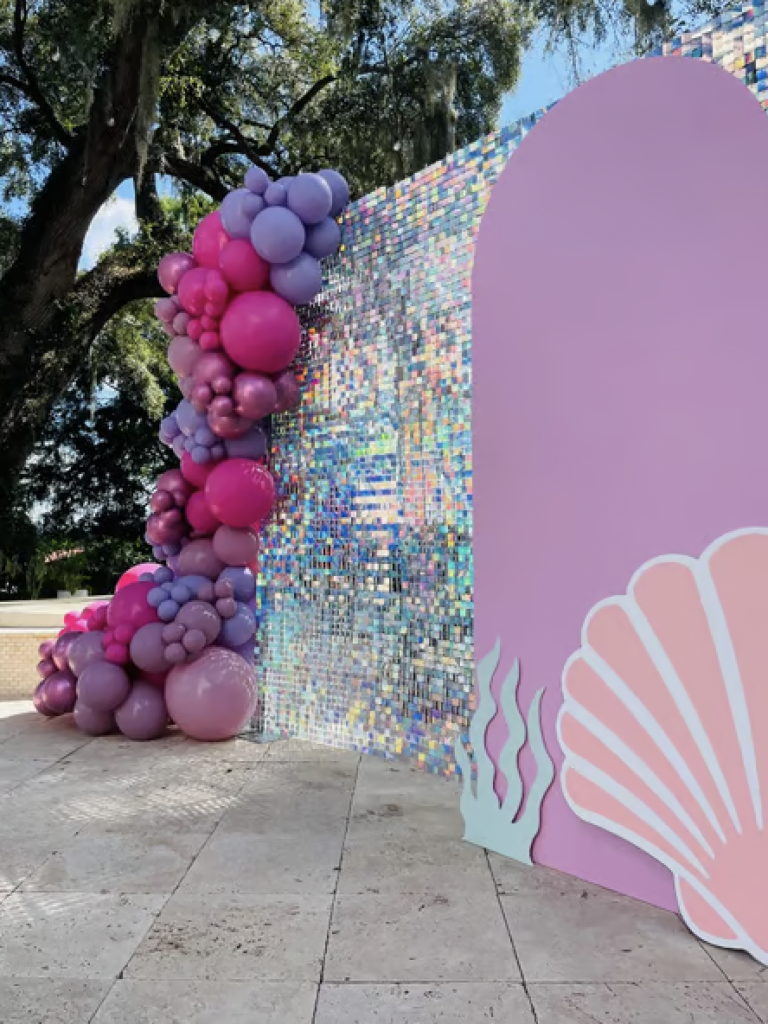 A pink wall and silver shimmer wall decorated with shells and purple/pink balloons for a mermaid themed birthday party. 