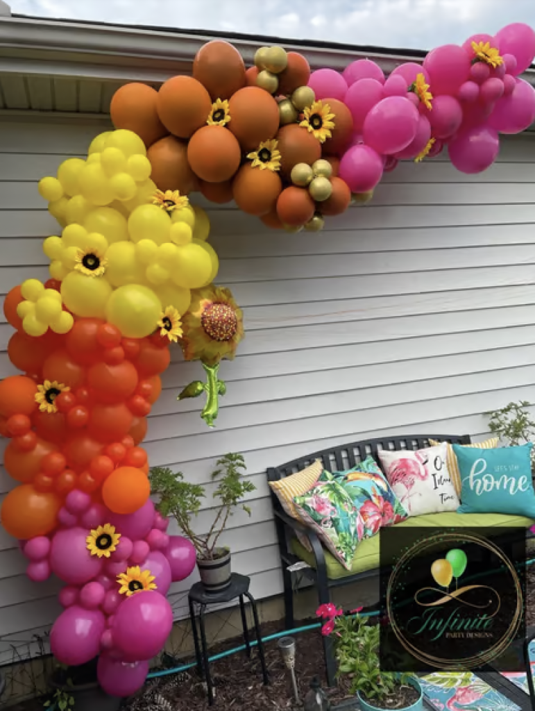 An orange, pink and yellow balloon garland decorated with sunflowers for an outdoor event.  